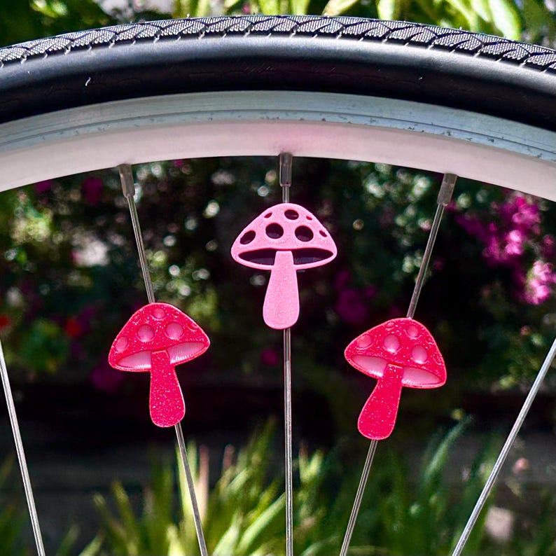 Puede incluir: Tres cuentas para radios en forma de seta de color rosa brillante fijadas a los radios met&aacute;licos de una rueda de bicicleta. Cada seta presenta una superficie texturizada y brillante con recortes circulares en el sombrero.