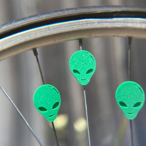 May include: Close-up of a bicycle wheel with three green alien head-shaped decorations attached to the spokes. The alien heads have large oval eyes and a small smile. The wheel rim is silver.