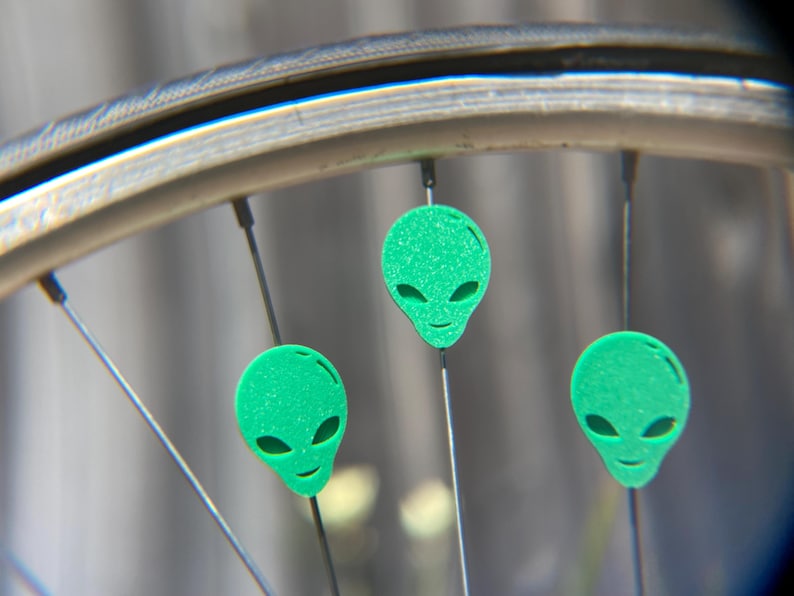 May include: Close-up of a bicycle wheel with three green alien head-shaped decorations attached to the spokes. The alien heads have large oval eyes and a small smile. The wheel rim is silver.