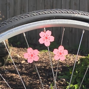 May include: Close-up of a bicycle wheel with three pink cherry blossom decorations attached to the spokes. The blossoms have a red center and are set against a backdrop of a black tire and a wooden fence.