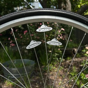 May include: A bicycle wheel with three silver UFO-shaped spoke decorations. The decorations are detailed with lines and are attached to the spokes. The tire is black, and the wheel is set against a blurred green background.