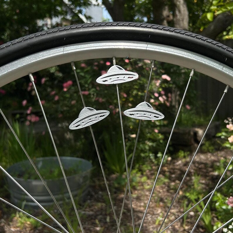 May include: A bicycle wheel with three silver UFO-shaped spoke decorations. The decorations are detailed with lines and are attached to the spokes. The tire is black, and the wheel is set against a blurred green background.