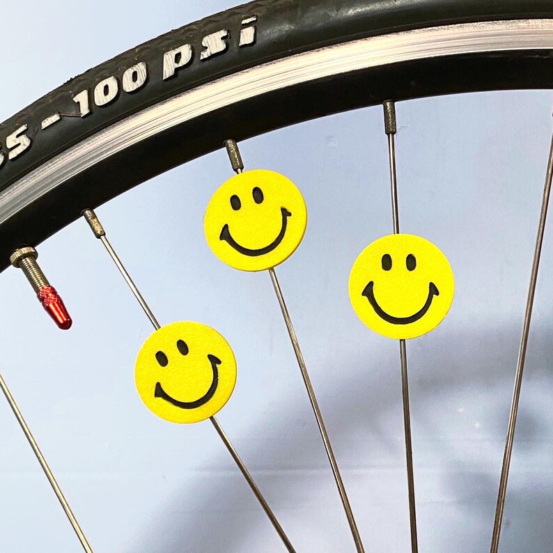 May include: Close-up of a bicycle wheel with three yellow smiley face spoke decorations. The tire is black with white text that reads "5 - 100 PSI". The valve stem has a red cap.