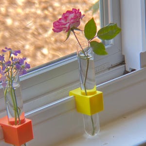 May include: Two glass bud vases, one with a pink and white rose and the other with purple flowers, sit in yellow and orange holders on a windowsill. The vases are filled with water, and the window frame is white.