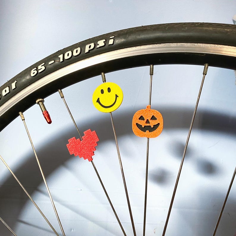 May include: A bicycle wheel with decorative spoke charms. The charms include a yellow smiley face, a red pixelated heart, and an orange jack-o'-lantern. The tire has the text "65-100 psi".