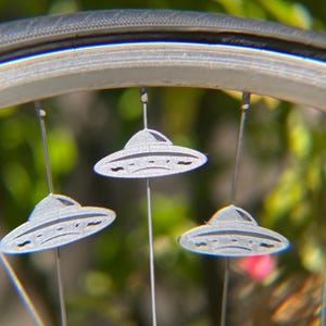 May include: Close-up of a bicycle wheel with three silver, UFO-shaped ornaments hanging from the spokes. The ornaments have a detailed design and are suspended by thin wires. The background is blurred with green and pink hues.