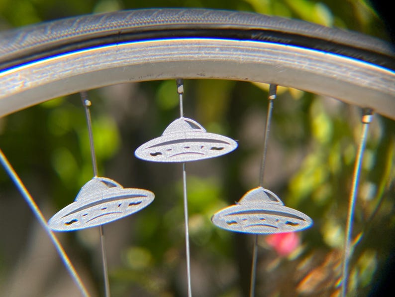 May include: Close-up of a bicycle wheel with three silver, UFO-shaped ornaments hanging from the spokes. The ornaments have a detailed design and are suspended by thin wires. The background is blurred with green and pink hues.