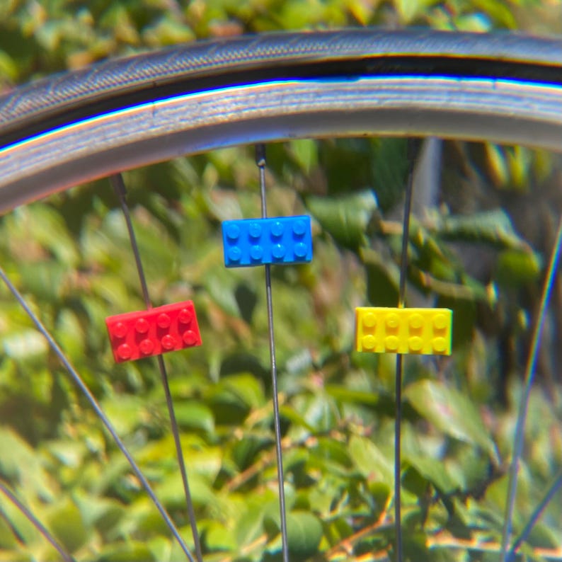 May include: A bicycle wheel with three colorful toy building blocks attached to the spokes. The blocks are red, blue, and yellow. The background is a blurred view of green foliage.