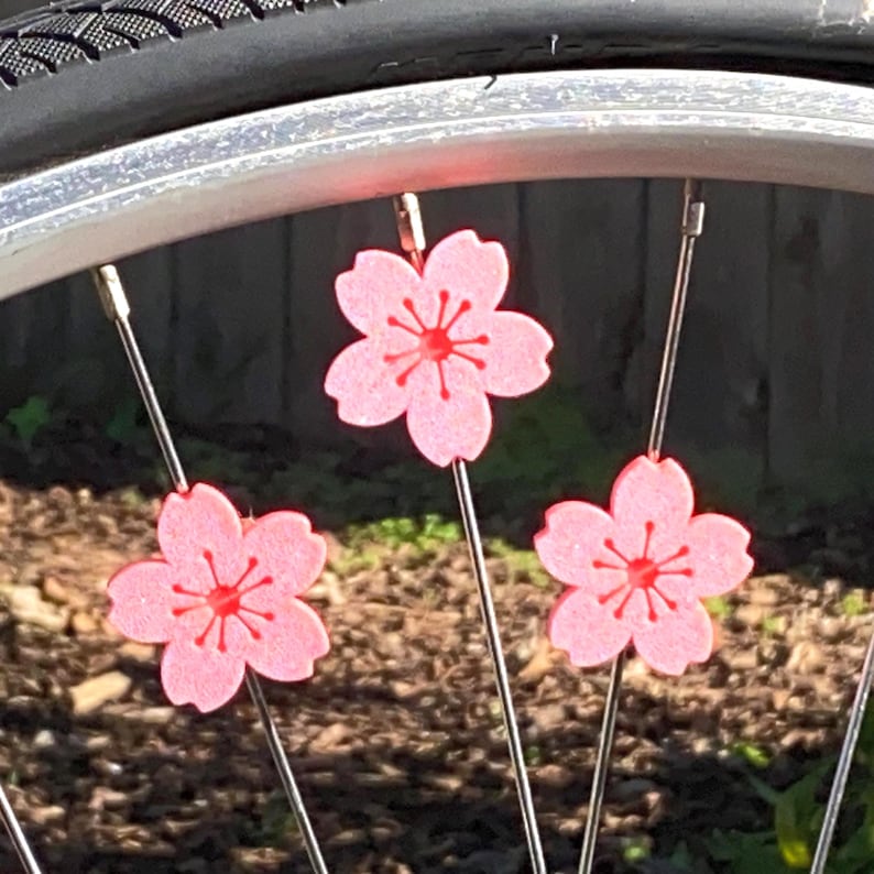 May include: Pink cherry blossom wheel spoke decorations. Three pink flower-shaped decorations with red centers are attached to the spokes of a bicycle wheel. The wheel is silver, and the tire is black.