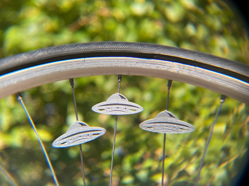 May include: Close-up of a bicycle wheel with three silver UFO-shaped charms attached to the spokes. The tire is black, and the background is a blurred green.