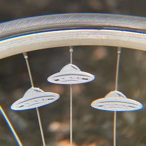 May include: Close-up of a bicycle wheel with three silver UFO-shaped spoke decorations. The wheel rim is silver, and the spokes are thin and metallic. The background is blurred, suggesting an outdoor setting.