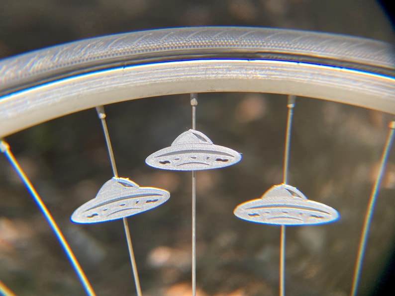 May include: Close-up of a bicycle wheel with three silver UFO-shaped spoke decorations. The wheel rim is silver, and the spokes are thin and metallic. The background is blurred, suggesting an outdoor setting.