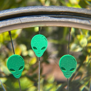 May include: Close-up of a bicycle wheel with three green alien-shaped ornaments attached to the spokes. The ornaments have oval eyes and a smiling mouth. The background is blurred green foliage.