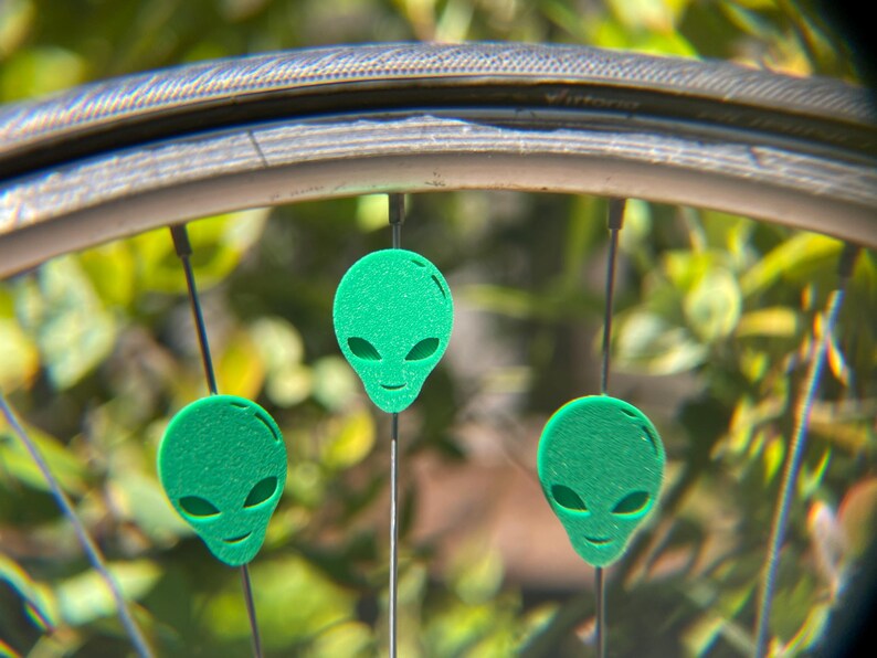 May include: Close-up of a bicycle wheel with three green alien-shaped ornaments attached to the spokes. The ornaments have oval eyes and a smiling mouth. The background is blurred green foliage.