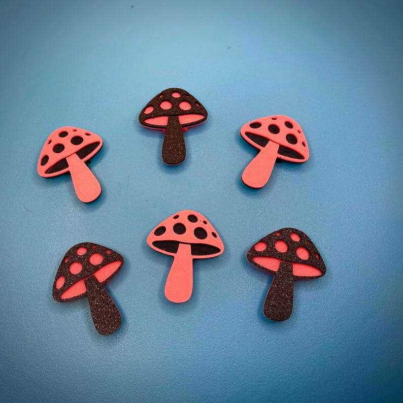 Puede incluir: Un conjunto de cinco dijes con forma de hongo dispuestos en un patr&oacute;n circular. Tres dijes presentan un sombrero negro brillante con puntos rosas y un tallo rosa s&oacute;lido, mientras que dos dijes tienen un sombrero rosa s&oacute;lido con puntos negros.