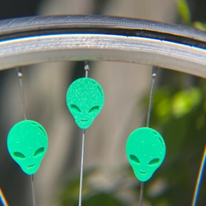 May include: Close-up of a bicycle wheel with three green alien-shaped reflectors attached to the spokes. The reflectors have a glittery texture and a smiling face design. The wheel rim is silver.