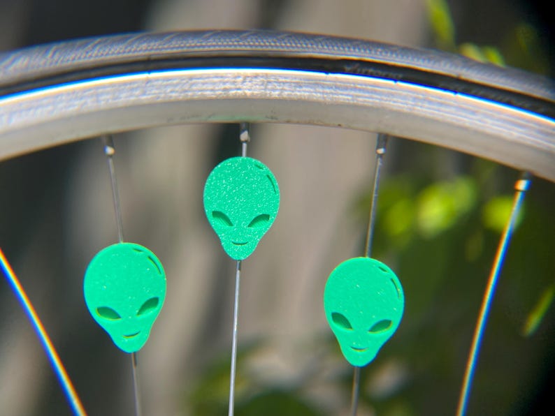 May include: Close-up of a bicycle wheel with three green alien-shaped reflectors attached to the spokes. The reflectors have a glittery texture and a smiling face design. The wheel rim is silver.