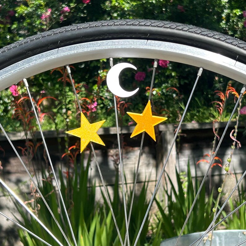 May include: A bicycle wheel with a black tire and silver spokes. The wheel is decorated with a white crescent moon and two yellow star-shaped charms. The background features green plants and flowers.