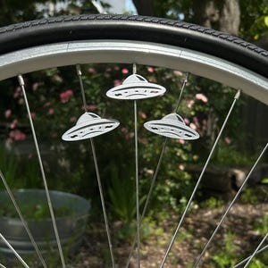 Könnte beinhalten: Nahaufnahme eines Fahrradreifens mit drei silbernen, UFO-förmigen Reflektoren an den Speichen. Die Reflektoren haben ein detailliertes Design. Der Hintergrund ist verschwommen mit grünem Laub und rosa Blüten.