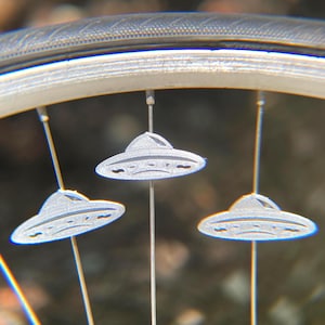 May include: Close-up of a bicycle wheel with three silver UFO-shaped spoke decorations. The decorations are suspended from the spokes and are detailed with etched lines. The tire and rim are visible in the background.