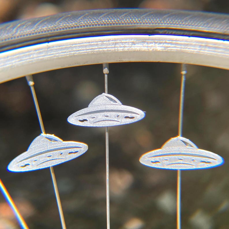 May include: Close-up of a bicycle wheel with three silver UFO-shaped spoke decorations. The decorations are suspended from the spokes and are detailed with etched lines. The tire and rim are visible in the background.