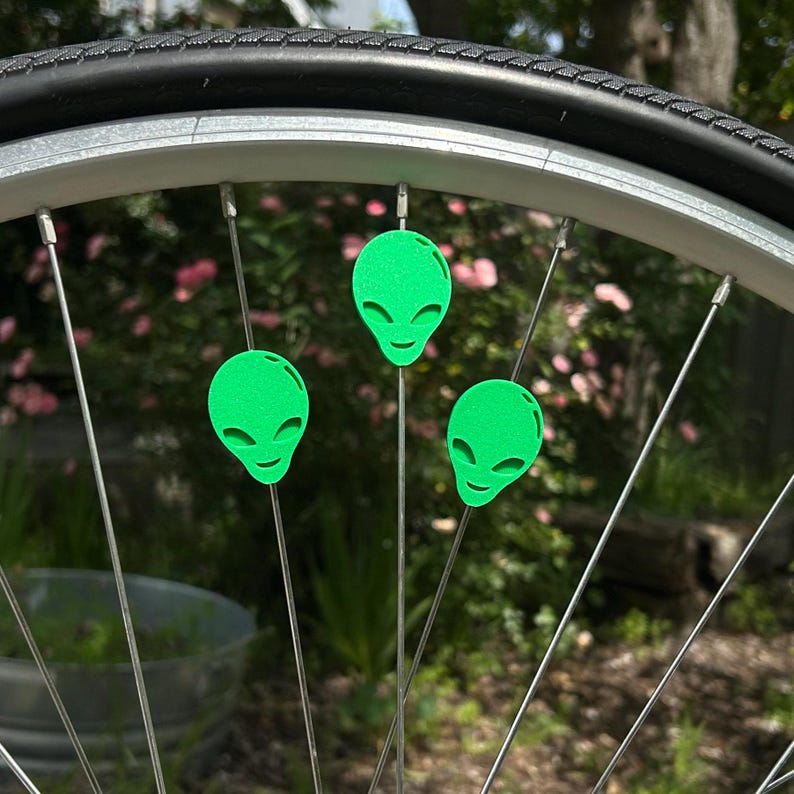 May include: A bicycle wheel with three neon green alien head spoke decorations. The alien heads have large eyes and a small smile. The wheel has a black tire and silver spokes.
