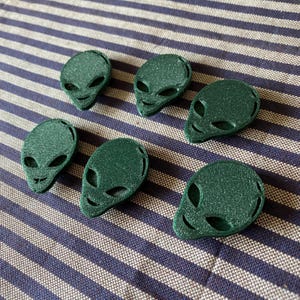 May include: Six green alien head-shaped objects with large oval eyes and small smiles are arranged on a blue and white striped surface. The objects have a textured, glittery appearance, suggesting a 3D-printed material.