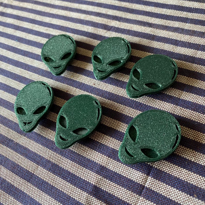 May include: Six green alien head-shaped objects with large oval eyes and small smiles are arranged on a blue and white striped surface. The objects have a textured, glittery appearance, suggesting a 3D-printed material.