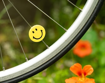 May include: A bicycle wheel with a yellow smiley face sticker attached to a spoke. The wheel has a black tire and silver spokes. An orange flower is in the background.