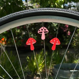 Puede incluir: Tres peque&ntilde;as decoraciones con forma de seta sujetas a los radios met&aacute;licos de una rueda de bicicleta. La seta superior es de color rosa claro con recortes oscuros, mientras que las dos inferiores son de color rojo brillante y texturizadas.