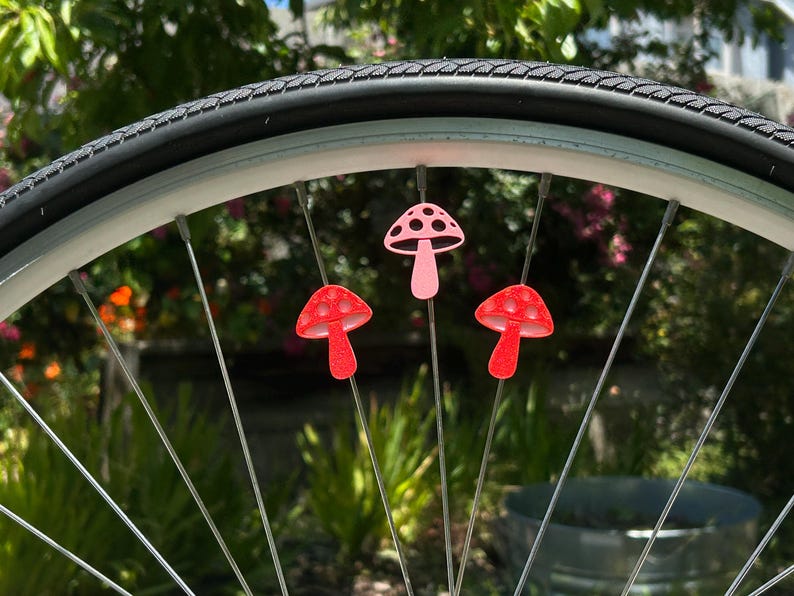 Puede incluir: Tres peque&ntilde;as decoraciones con forma de seta sujetas a los radios met&aacute;licos de una rueda de bicicleta. La seta superior es de color rosa claro con recortes oscuros, mientras que las dos inferiores son de color rojo brillante y texturizadas.