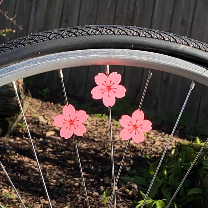 May include: Close-up of a bicycle wheel with three pink cherry blossom decorations attached to the spokes. The blossoms have a red center and are set against a backdrop of greenery and sunlight.
