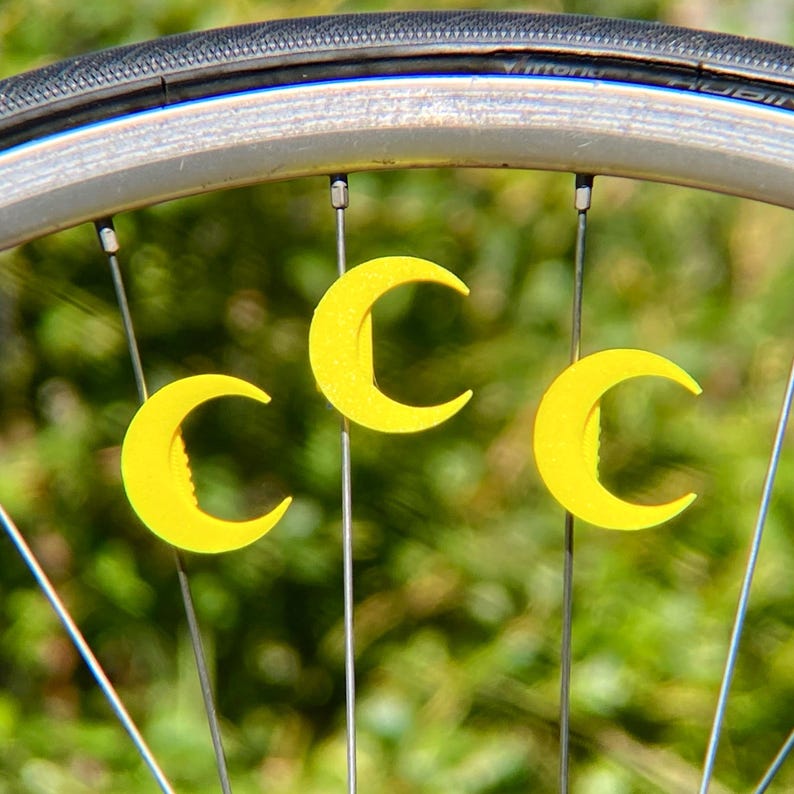 May include: Close-up of a bicycle wheel with three yellow crescent moon-shaped decorations attached to the spokes. The tire is black, and the wheel rim is silver. The background is a blurred green.