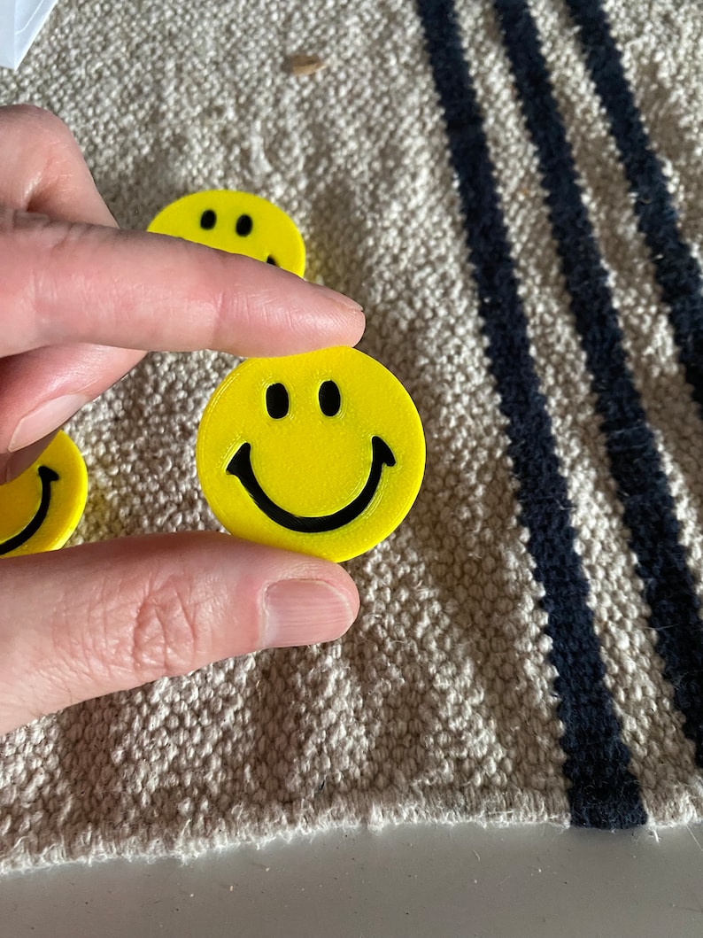 May include: Yellow smiley face with black eyes and a curved mouth. The smiley face is held between fingers. Several other smiley faces are visible in the background. The smiley faces are on a textured, beige surface.