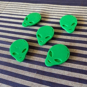 May include: Five green alien head-shaped objects with large oval eyes and small smiles. The objects are arranged on a blue and white striped surface. The objects are a solid, matte green color.
