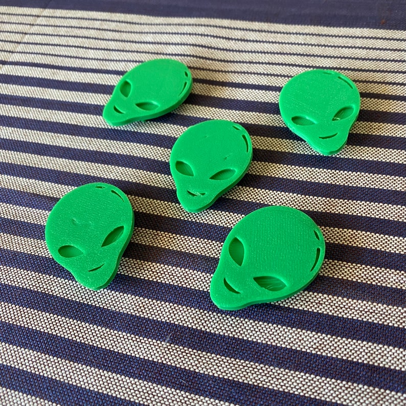 May include: Five green alien head-shaped objects with large oval eyes and small smiles. The objects are arranged on a blue and white striped surface. The objects are a solid, matte green color.