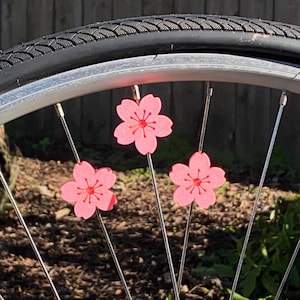May include: Close-up of a bicycle wheel with three pink cherry blossom decorations attached to the spokes. The blossoms have a red center and are set against a backdrop of a black tire and a blurred green and brown background.