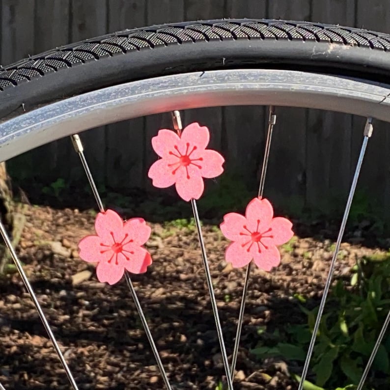May include: Close-up of a bicycle wheel with three pink cherry blossom decorations attached to the spokes. The blossoms have a red center and are set against a backdrop of a black tire and a blurred green and brown background.