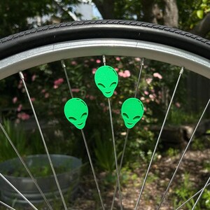 May include: Close-up of a bicycle wheel with three neon green alien head spoke decorations. The decorations are attached to the silver spokes of the wheel. The tire is black, and the background is blurred with greenery.