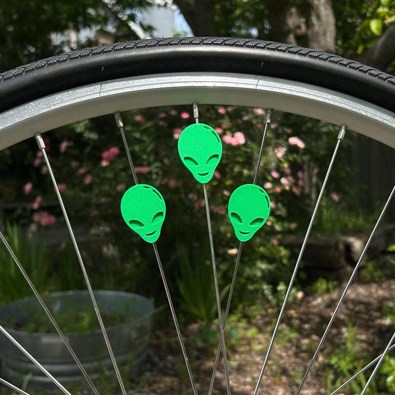 May include: Close-up of a bicycle wheel with three neon green alien head spoke decorations. The decorations are attached to the silver spokes of the wheel. The tire is black, and the background is blurred with greenery.