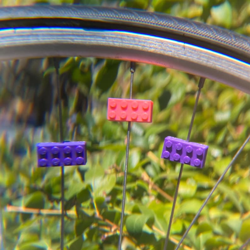 May include: Close-up of a bicycle wheel with three colorful Lego-style brick decorations attached to the spokes. The bricks are pink and purple, adding a playful touch to the bike.
