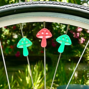 Puede incluir: Tres cuentas para radios en forma de seta fijadas a los radios de una rueda de bicicleta. Las dos cuentas exteriores son de color verde brillante y la central es de color rojo brillante. Cada cuenta tiene un sombrero redondeado.