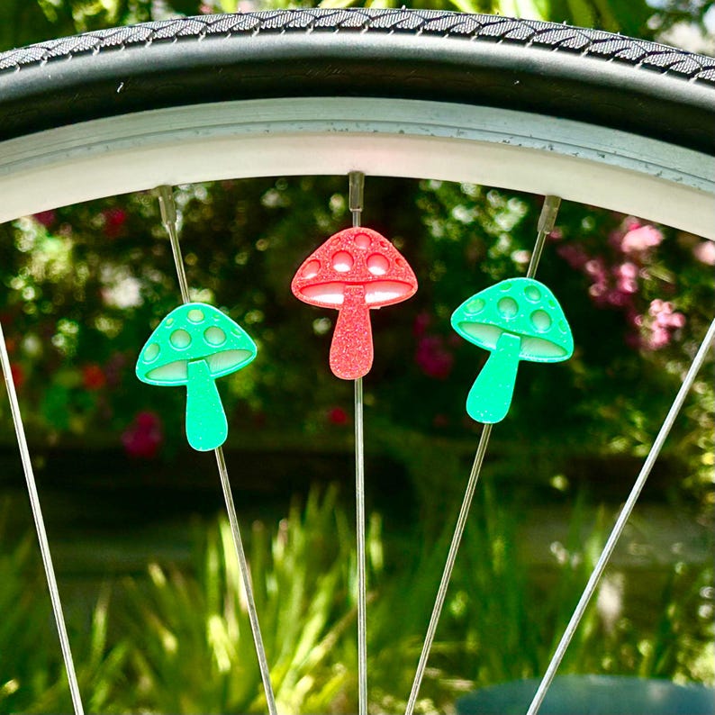 Puede incluir: Tres cuentas para radios en forma de seta fijadas a los radios de una rueda de bicicleta. Las dos cuentas exteriores son de color verde brillante y la central es de color rojo brillante. Cada cuenta tiene un sombrero redondeado.