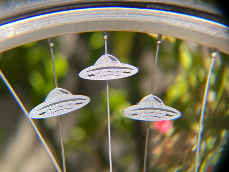 May include: Close-up of a bicycle wheel with three silver UFO-shaped ornaments hanging from the spokes. The ornaments have a detailed design and are suspended by thin wires. The background is blurred with green and pink hues.