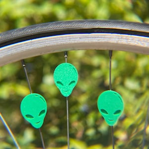 May include: Close-up of a bicycle wheel with three green alien head-shaped spoke decorations. The decorations are evenly spaced and attached to the spokes. The background is blurred green foliage.
