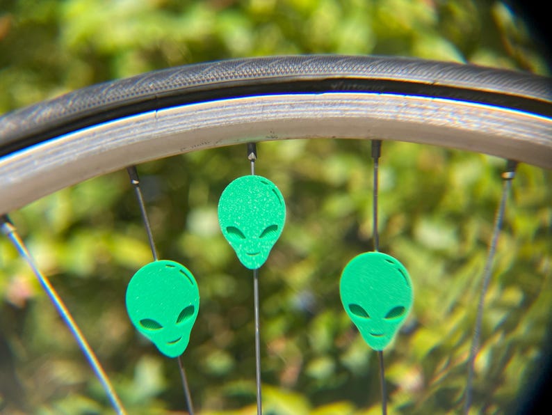 May include: Close-up of a bicycle wheel with three green alien head-shaped spoke decorations. The decorations are evenly spaced and attached to the spokes. The background is blurred green foliage.