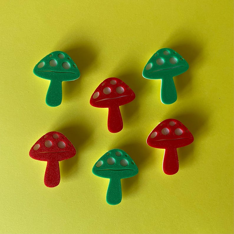Puede incluir: Una colecci&oacute;n de seis botones con forma de seta dispuestos en un patr&oacute;n. Tres botones son de color verde vibrante y tres son de color rojo brillante. Cada bot&oacute;n presenta un sombrero redondeado con tres hendiduras blancas y un tallo.