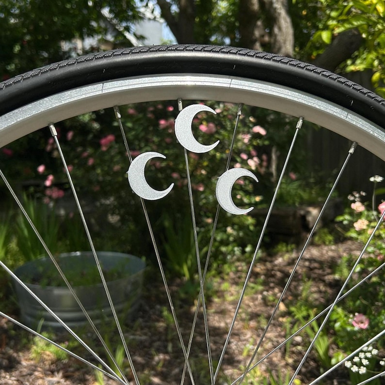 May include: Close-up of a bicycle wheel with silver spokes and a black tire. Three silver crescent moon charms are attached to the spokes. The background is blurred with green foliage and a metal container.