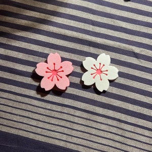 May include: Two flower-shaped embellishments. One is pink with a red center, and the other is white with a red center. Both have a five-petal design with a starburst pattern in the center. They are placed on a blue and white striped fabric.