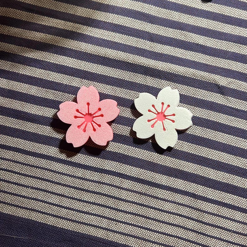 May include: Two flower-shaped embellishments. One is pink with a red center, and the other is white with a red center. Both have a five-petal design with a starburst pattern in the center. They are placed on a blue and white striped fabric.